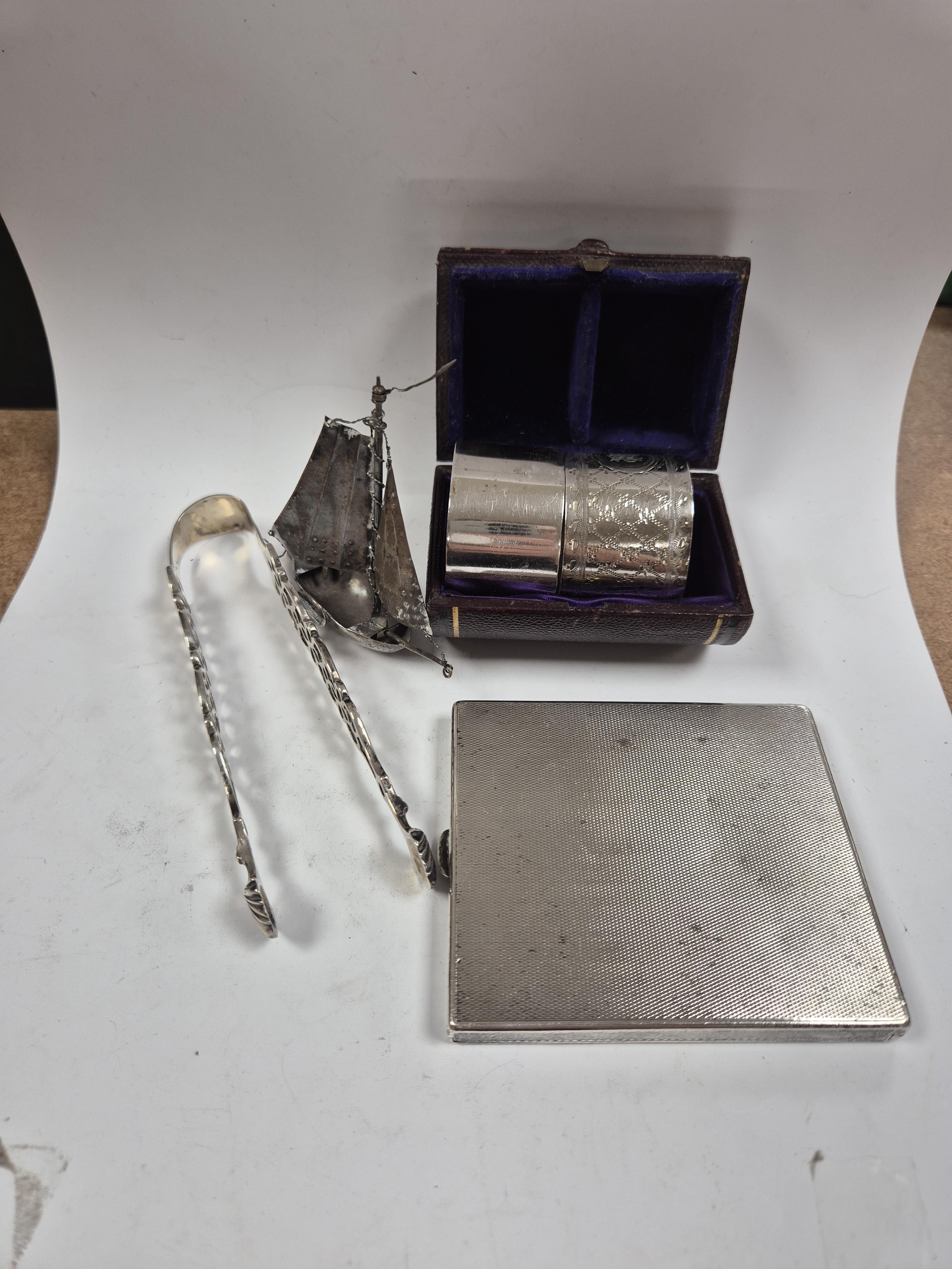 A 1930's engine turned silver cigarette case, 88mm, two silver napkin rings, a white metal miniature model of a boat and a pair of Georgian silver sugar tongs (a.f.). Condition - poor to fair